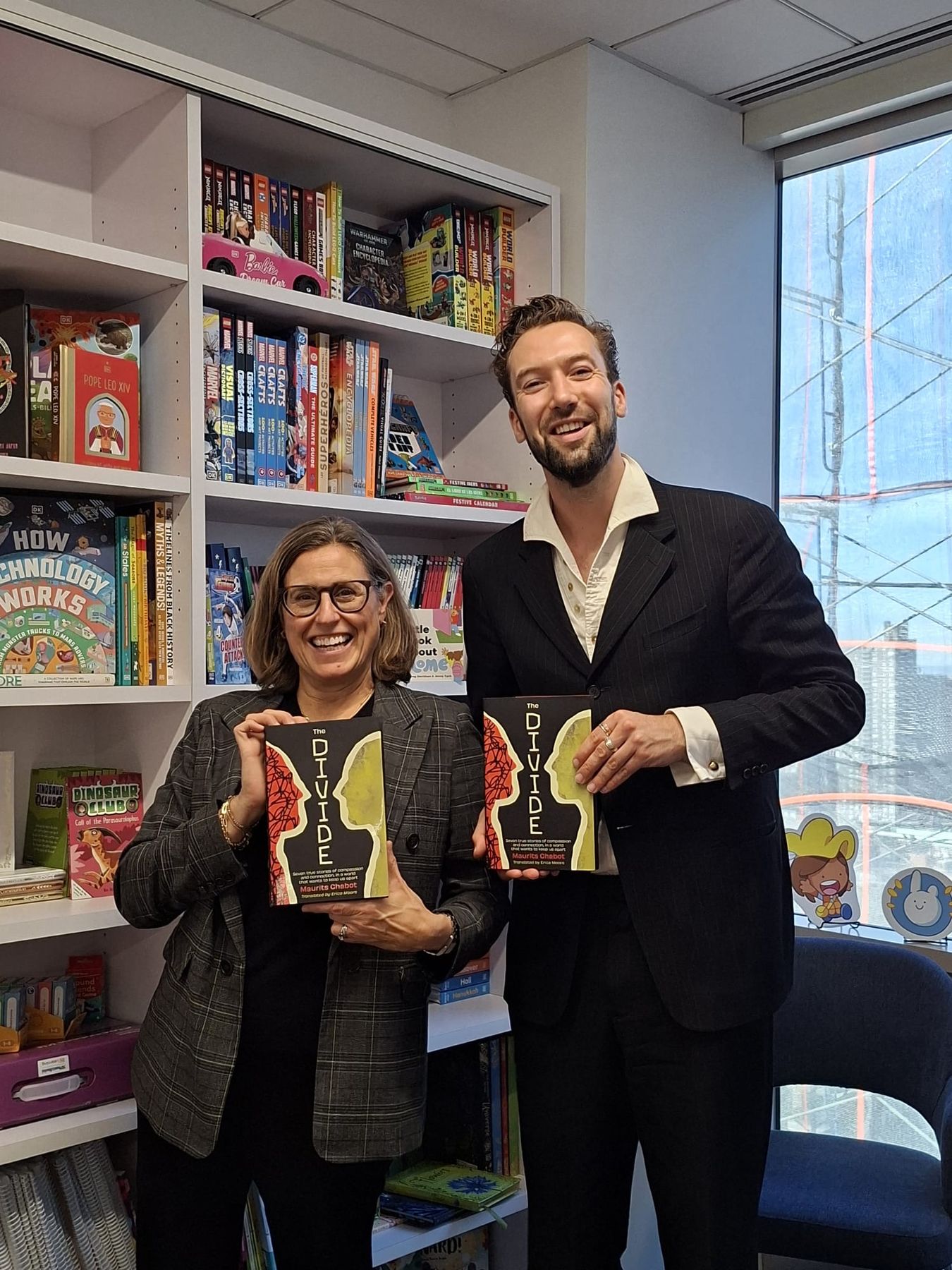 Maurits Chabot en een collega van de uitgeverij staan glimlachend naast elkaar en houden elk een exemplaar van The Divide vast, tussen de boekenplanken van een redactiekantoor.