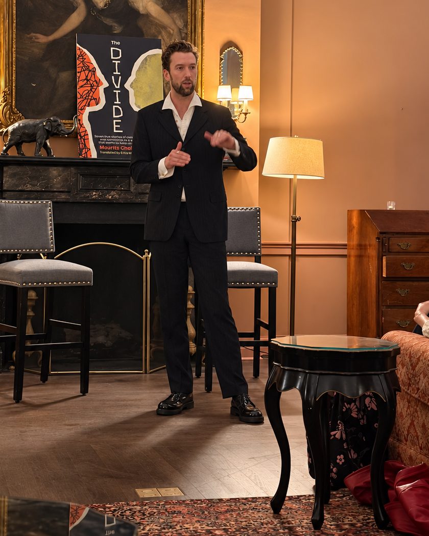 Een warm verlichte zaal met schouw, staande lampen en barkrukken; Maurits Chabot staat in het midden en vertelt, naast de omslag van The Divide.