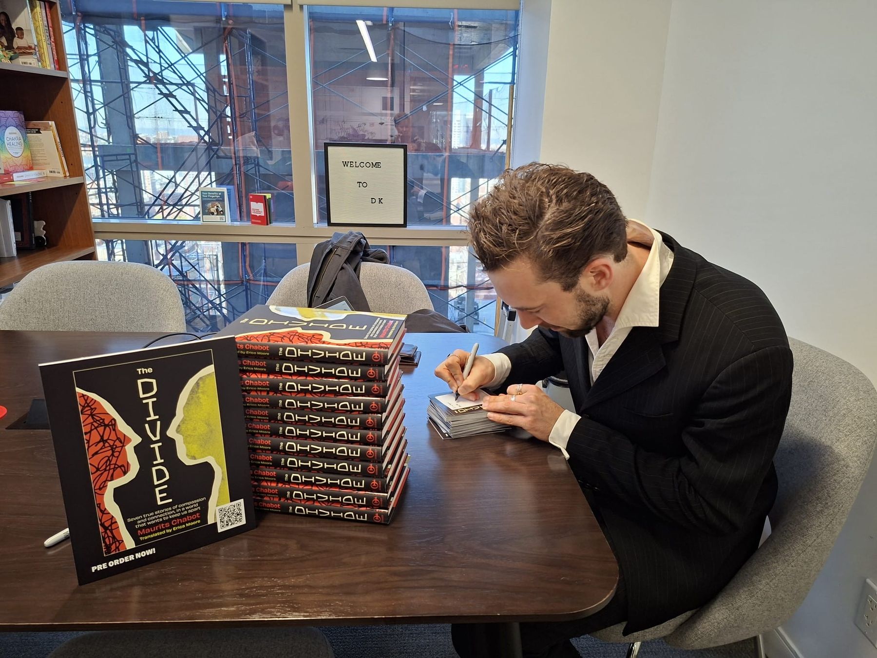 Maurits Chabot zit aan een vergadertafel en signeert boekplaten; naast hem staat een stapel exemplaren van The Divide en een opstaande kartonnen boekomslag.